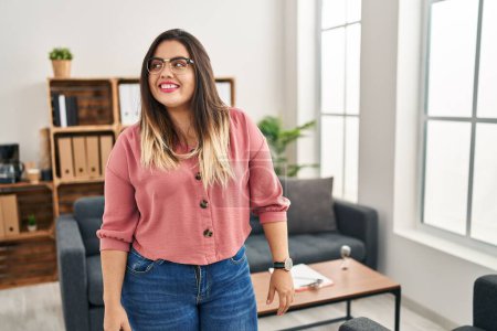 Téléchargez les photos : Young hispanic woman working at the office wearing glasses looking to side, relax profile pose with natural face and confident smile. - en image libre de droit