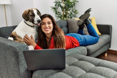 Photo for Young woman using laptop lying on sofa with dog at home - Royalty Free Image