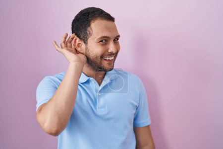 Foto de Hombre hispano de pie sobre fondo rosa sonriendo con la mano sobre el oído escuchando un rumor o chismes. concepto de sordera. - Imagen libre de derechos