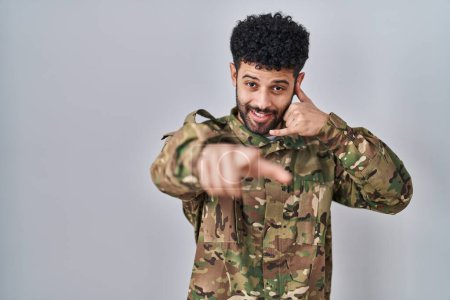 Photo for Arab man wearing camouflage army uniform smiling doing talking on the telephone gesture and pointing to you. call me. - Royalty Free Image