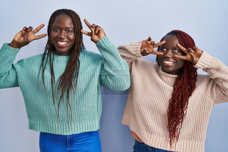 Foto de Dos mujeres africanas de pie sobre fondo azul haciendo símbolo de paz con los dedos sobre la cara, sonriendo alegre mostrando la victoria - Imagen libre de derechos