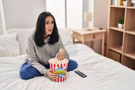Foto de Mujer hispana comiendo palomitas de maíz viendo una película en la cama sin pistas y expresión confusa. concepto de duda. - Imagen libre de derechos