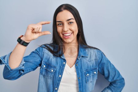 Foto de Mujer hispana de pie sobre fondo azul sonriente y seguro gesto con la mano haciendo signo de tamaño pequeño con los dedos mirando y la cámara. concepto de medida. - Imagen libre de derechos