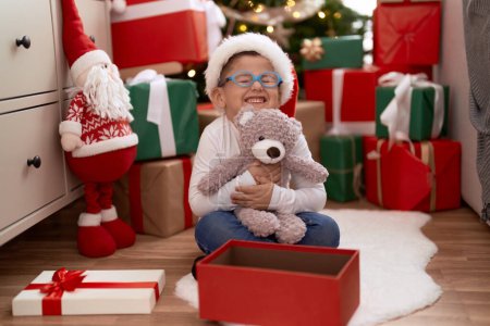Foto de Adorable niño hispano abrazando oso de peluche sentado en el suelo por regalos de Navidad en casa - Imagen libre de derechos