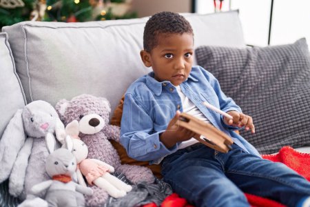 Photo for African american boy playing tambourine sitting on sofa by christmas tree at home - Royalty Free Image