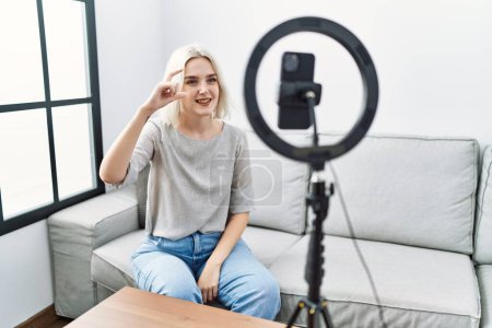 Téléchargez les photos : Young caucasian woman recording vlog tutorial with smartphone at home smiling and confident gesturing with hand doing small size sign with fingers looking and the camera. measure concept. - en image libre de droit