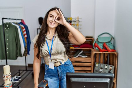 Téléchargez les photos : Jeune femme brune travaillant comme gestionnaire à la boutique de détail souriant heureux faire ok signe avec la main sur les yeux regardant à travers les doigts - en image libre de droit