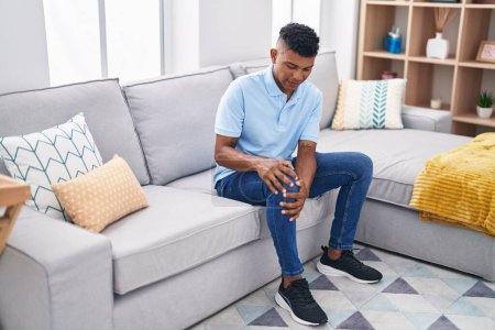 Téléchargez les photos : Jeune homme latino souffrant d'une blessure au genou assis sur le canapé à la maison - en image libre de droit
