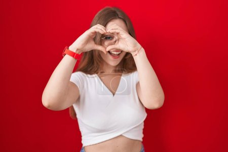 Téléchargez les photos : Jeune femme caucasienne debout sur fond rouge faisant forme de coeur avec la main et les doigts souriant en regardant à travers signe - en image libre de droit