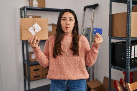 Foto de Young latin woman working at small business ecommerce holding credit card making fish face with mouth and squinting eyes, crazy and comical. - Imagen libre de derechos