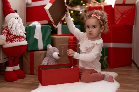 Foto de Adorable chica caucásica desembalaje regalo sentado en el suelo por árbol de Navidad en casa - Imagen libre de derechos