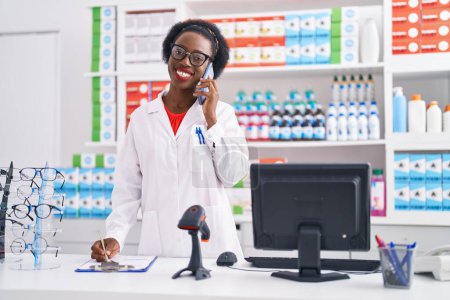 Foto de African american woman pharmacist talking on smartphone writing on document at pharmacy - Imagen libre de derechos