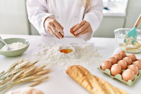 Foto de Young woman wearing cook uniform cracking egg on flour at kitchen - Imagen libre de derechos