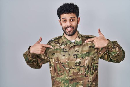 Foto de Hombre árabe vistiendo camuflaje uniforme del ejército mirando confiado con sonrisa en la cara, señalándose con los dedos orgullosos y felices. - Imagen libre de derechos