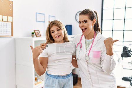 Photo for Pediatrician woman working at the clinic with little girl pointing thumb up to the side smiling happy with open mouth - Royalty Free Image