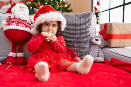 Photo for Adorable hispanic girl sucking stair decoration sitting on sofa by christmas tree at home - Royalty Free Image