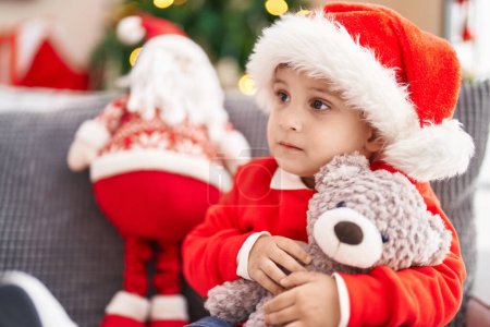 Photo for Adorable hispanic toddler hugging teddy bear sitting on sofa by christmas tree at home - Royalty Free Image