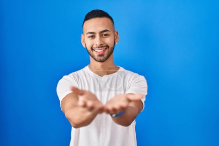 Téléchargez les photos : Young hispanic man standing over blue background smiling with hands palms together receiving or giving gesture. hold and protection - en image libre de droit