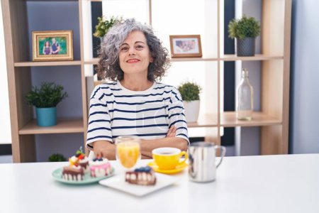 Téléchargez les photos : Middle age woman with grey hair eating pastries and drinking coffee for breakfast happy face smiling with crossed arms looking at the camera. positive person. - en image libre de droit
