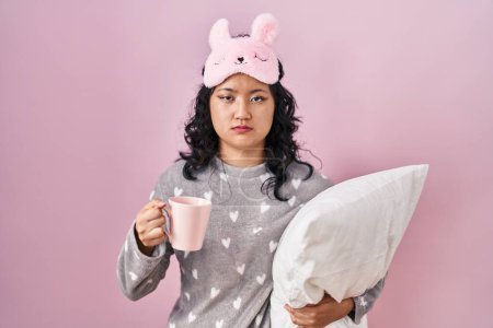 Photo for Young asian woman wearing sleep mask and pajama hugging pillow drinking coffee skeptic and nervous, frowning upset because of problem. negative person. - Royalty Free Image