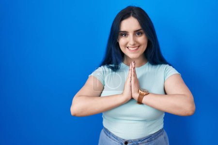 Foto de Young modern girl with blue hair standing over blue background praying with hands together asking for forgiveness smiling confident. - Imagen libre de derechos