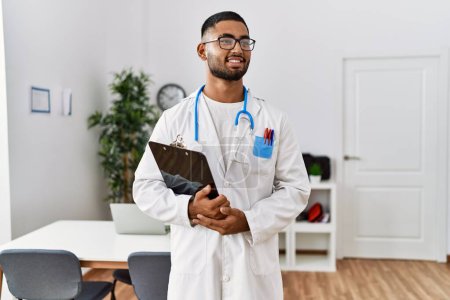 Photo for Young arab man wearing doctor uniform holding clipboard at clinic - Royalty Free Image
