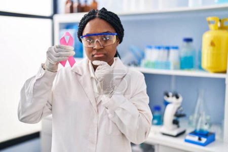Photo for African american woman working at scientist laboratory holding pink ribbon serious face thinking about question with hand on chin, thoughtful about confusing idea - Royalty Free Image