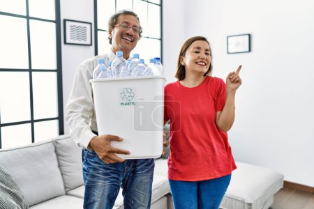 Téléchargez les photos : Couple interracial moyen âge tenant poubelle de recyclage avec des bouteilles en plastique à la maison souriant heureux pointant avec la main et le doigt sur le côté - en image libre de droit