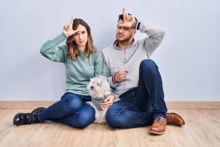 Foto de Young hispanic couple sitting on the floor with dog making fun of people with fingers on forehead doing loser gesture mocking and insulting. - Imagen libre de derechos