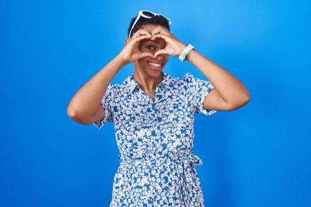 Foto de Mujer afroamericana de pie sobre fondo azul haciendo forma de corazón con la mano y los dedos sonriendo mirando a través de signo - Imagen libre de derechos