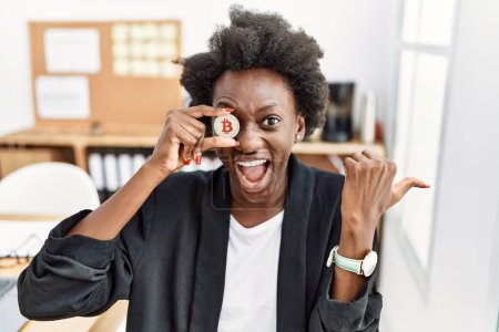 Téléchargez les photos : Africaine jeune femme tenant Bitcoin sur les yeux au bureau pointant pouce vers le côté souriant heureux avec bouche ouverte - en image libre de droit