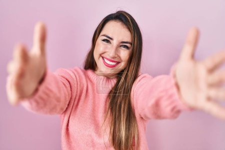 Foto de Mujer hispana joven de pie sobre fondo rosa mirando a la cámara sonriendo con los brazos abiertos para abrazarse. expresión alegre abrazando la felicidad. - Imagen libre de derechos