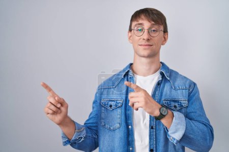 Foto de Caucasian blond man standing wearing glasses smiling and looking at the camera pointing with two hands and fingers to the side. - Imagen libre de derechos