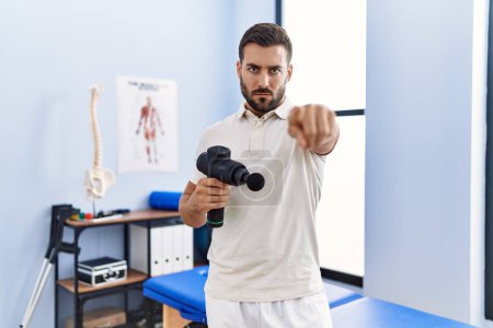 Photo for Handsome hispanic man holding therapy massage gun at physiotherapy center pointing with finger to the camera and to you, confident gesture looking serious - Royalty Free Image