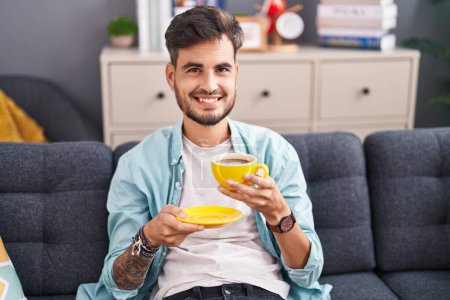 Photo for Young hispanic man drinking coffee sitting on sofa at home - Royalty Free Image
