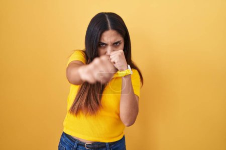 Téléchargez les photos : Jeune femme arabe debout sur fond jaune coups de poing poing pour se battre, attaque agressive et en colère, menace et violence - en image libre de droit