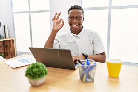 Téléchargez les photos : Jeune homme africain qui travaille au bureau en utilisant un ordinateur portable souriant positif faire ok signe avec la main et les doigts. expression réussie. - en image libre de droit