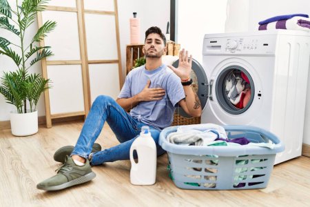 Photo for Young hispanic man putting dirty laundry into washing machine swearing with hand on chest and open palm, making a loyalty promise oath - Royalty Free Image