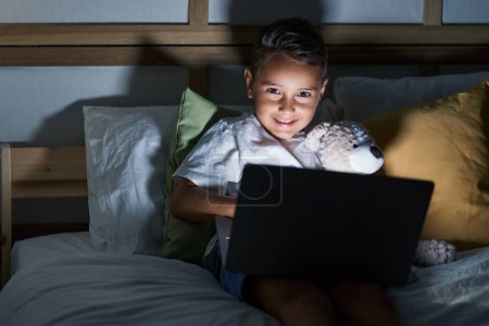 Foto de Adorable niño hispano usando portátil sentado en la cama en el dormitorio - Imagen libre de derechos