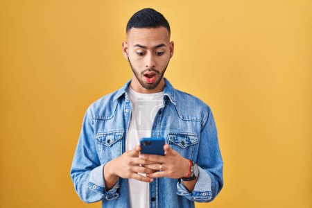 Téléchargez les photos : Young hispanic man using smartphone typing message in shock face, looking skeptical and sarcastic, surprised with open mouth - en image libre de droit