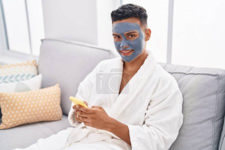 Photo for Young latin man relaxed on sofa with facial mask treatment using smartphone at home - Royalty Free Image