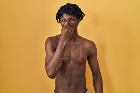 Foto de Young african man with dreadlocks standing shirtless smelling something stinky and disgusting, intolerable smell, holding breath with fingers on nose. bad smell - Imagen libre de derechos