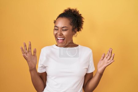 Photo for Young hispanic woman with curly hair standing over yellow background celebrating mad and crazy for success with arms raised and closed eyes screaming excited. winner concept - Royalty Free Image