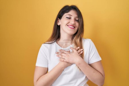 Photo for Young brunette woman standing over yellow background smiling with hands on chest with closed eyes and grateful gesture on face. health concept. - Royalty Free Image