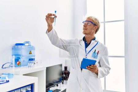 Photo for Young caucasian man wearing scientist uniform holding test tube and touchpad at laboratory - Royalty Free Image