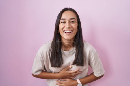 Foto de Joven mujer hispana de pie sobre fondo rosa sonriendo y riendo en voz alta porque divertida broma loca con las manos en el cuerpo. - Imagen libre de derechos