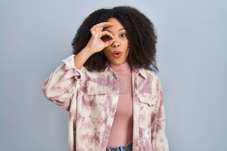 Foto de Young african american woman standing over blue background doing ok gesture shocked with surprised face, eye looking through fingers. unbelieving expression. - Imagen libre de derechos