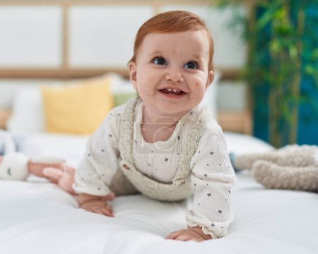 Foto de Adorable pelirroja niño sonriendo confiado arrastrándose en la cama en el dormitorio - Imagen libre de derechos