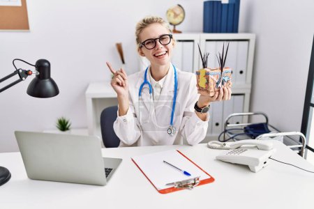 Photo for Young doctor woman holding model of human anatomical skin and hair at the clinic smiling happy pointing with hand and finger to the side - Royalty Free Image