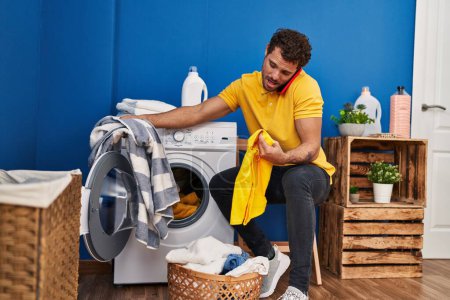 Téléchargez les photos : Young hispanic man talking on the smartphone washing clothes at laundry room - en image libre de droit